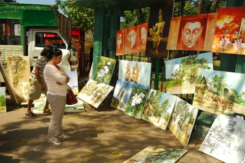 A woman examines various paintings displayed at an outdoor market, showcasing her interest in art and creativity.