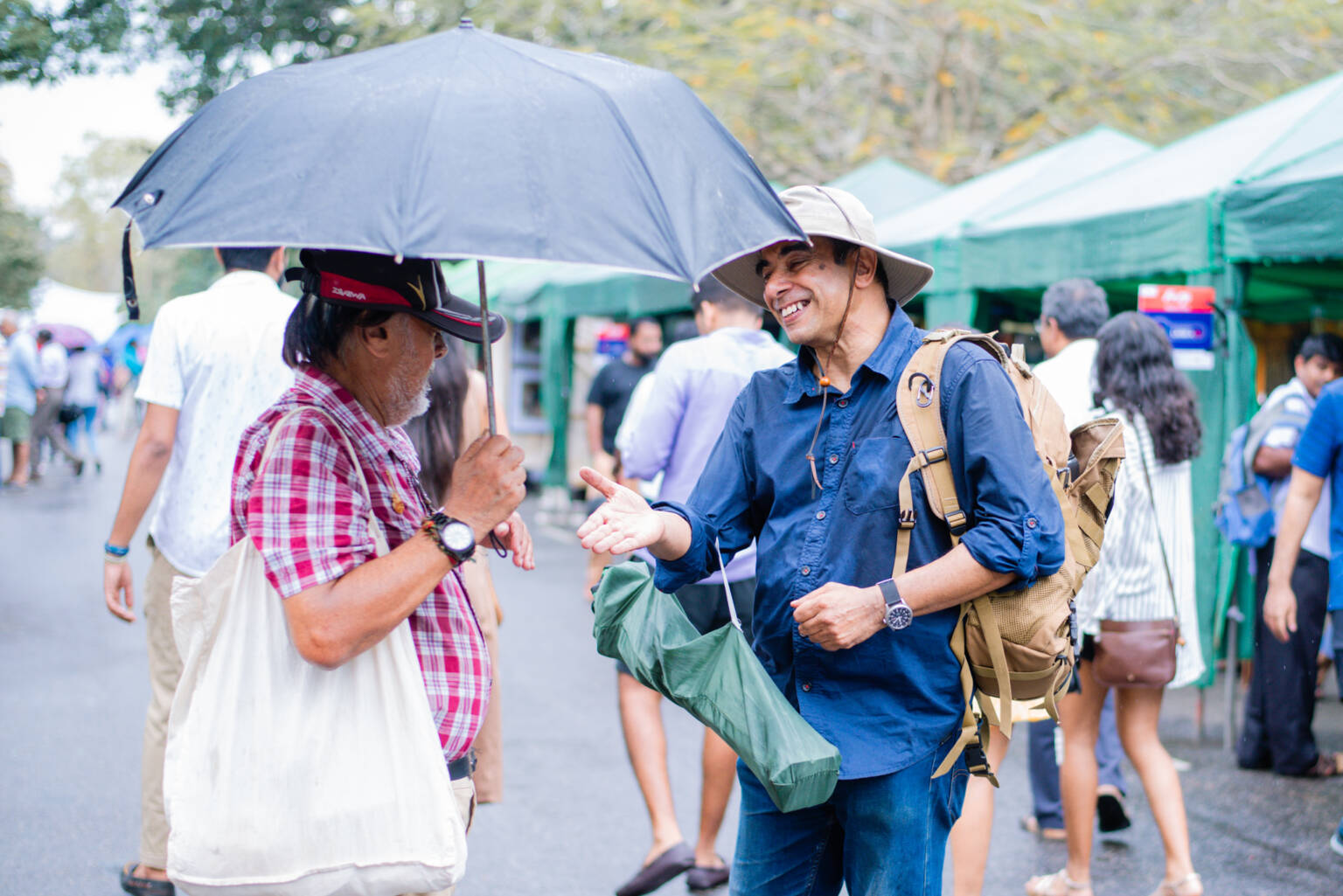 Two men engage in conversation, with one providing shelter from the rain by holding an umbrella over the other.