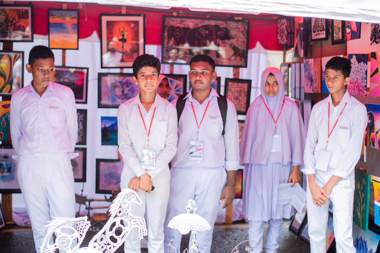 A group of school students stands under a tent displaying various pictures, engaged in conversation and activities.