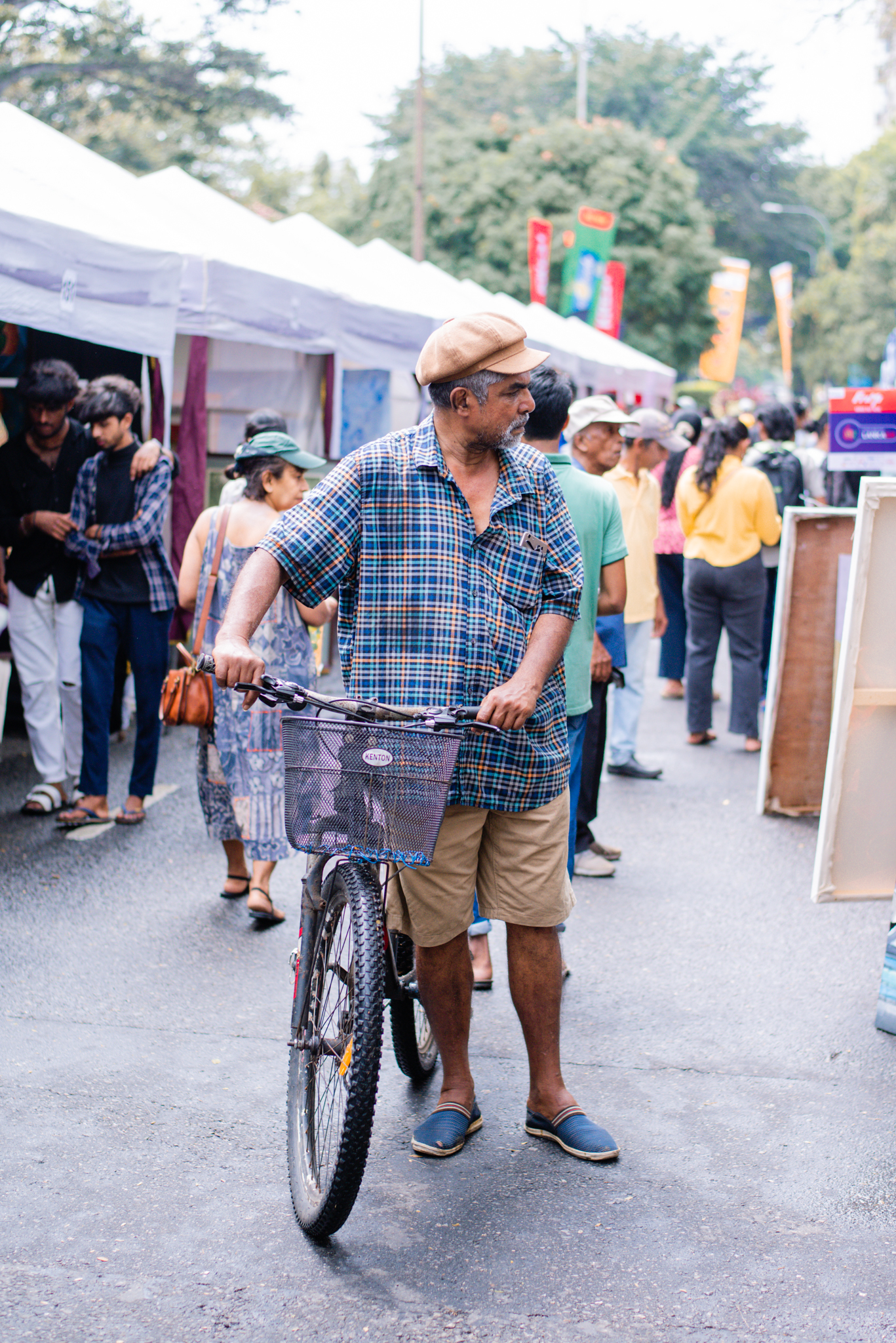 A man dressed in a blue shirt strolls with his bicycle, gazing at a painting display on the street.