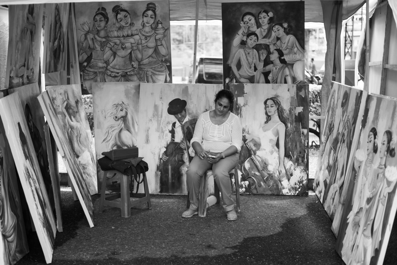 A man wearing a blue shirt walks beside his bicycle, observing a showcase of paintings on the street.